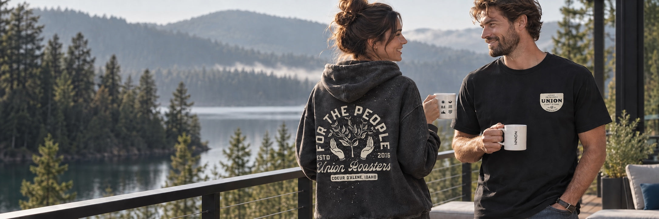 Two friends wearing Union clothing and holding mugs beside a Northwest lake